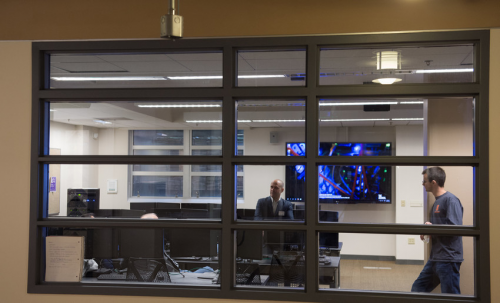 Hinds hall computer lab. View from outside looking in. Multiple computer stations. A person leans against a station. Another leans against a wall in the computer lab,