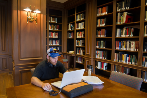View of Safire room in Bird Library. A student seated at a large table with laptop. Behind him is a library.