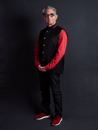 Dr. Deepak Chopra wearing a red shirt, black vest, jeans and red sneakers.