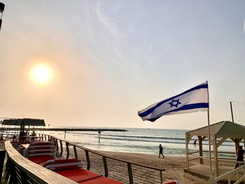 The Israeli flag flies over the coastline at sunset.