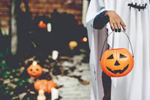A person in a bed sheet ghost costume holds out a jack-o-lantern bucket for candy.