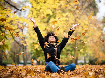 A person sits cross legged among yellow leaves, throwing leaves into the air. 
