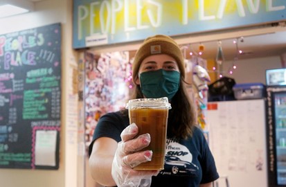 A person in a beanie and green facemask holds out an iced coffee at Peoples Place.