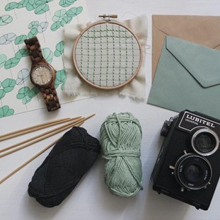 A tablescape of yarn, knitting needles, an embroidery hoop and film camera