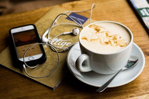 A phone with headphones rests by a cappuccino 