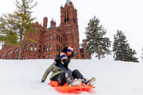 Two students on a bright orange sled sledding down the hill in front of Crouse College on a snowy winter day.