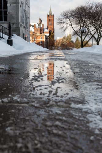 A closeup of snow and sidewalk slush outside the Hall of Languages with Crouse college reflected in the slush puddle. 