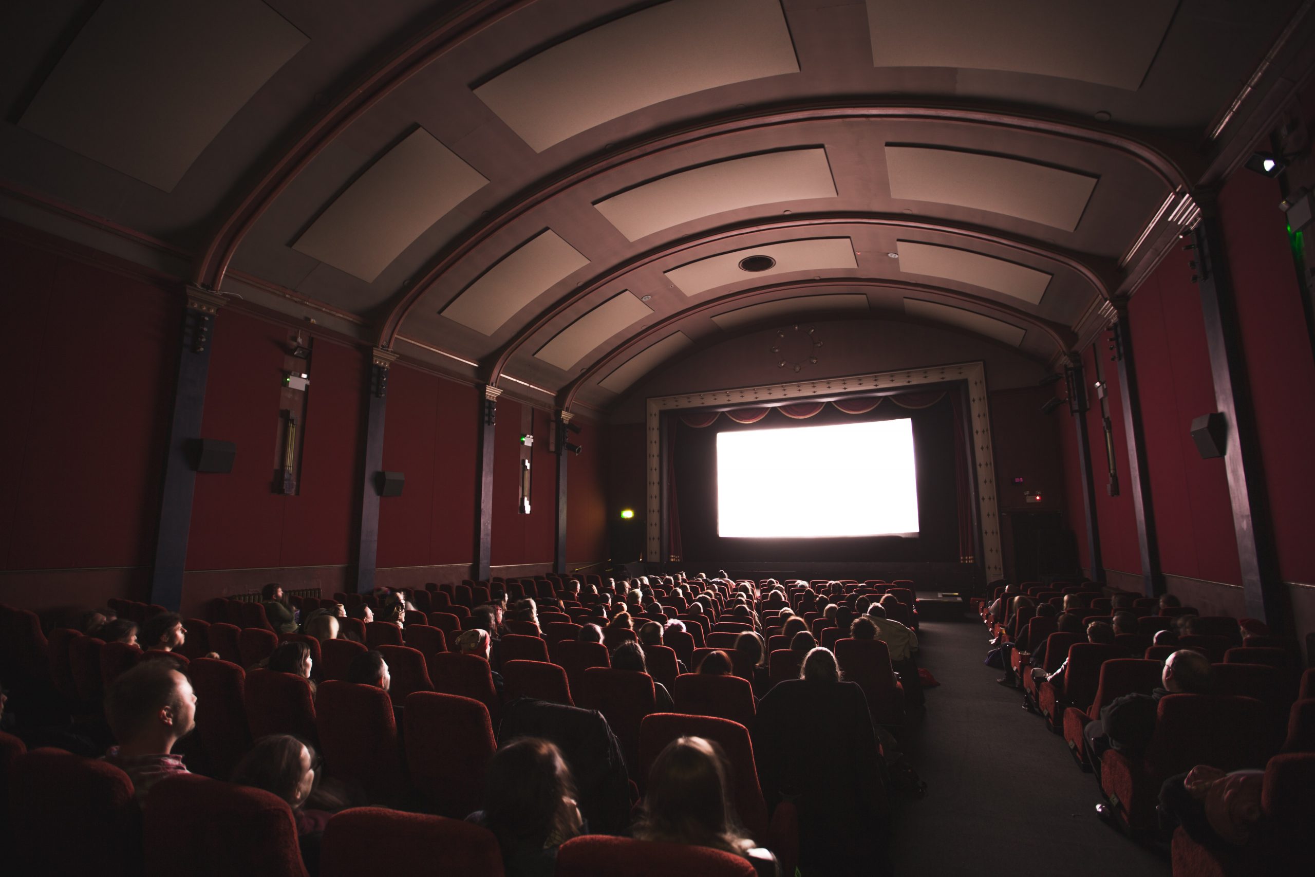 A crowd sits in a movie theater waiting for a movie to start. Movie screen is lit up white.