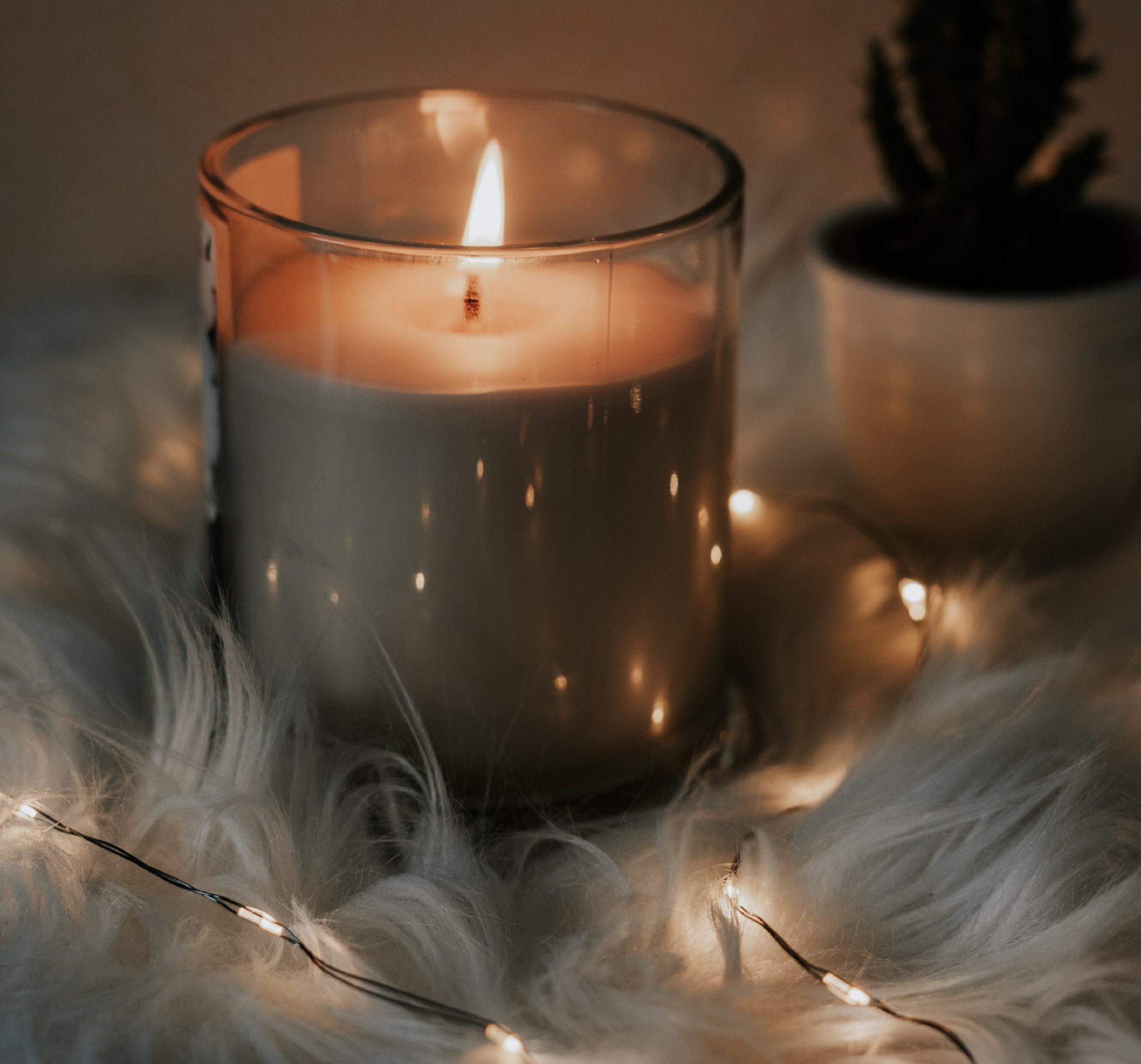 A lit candle sits on a fuzzy blanket surrounded by a string of lights.