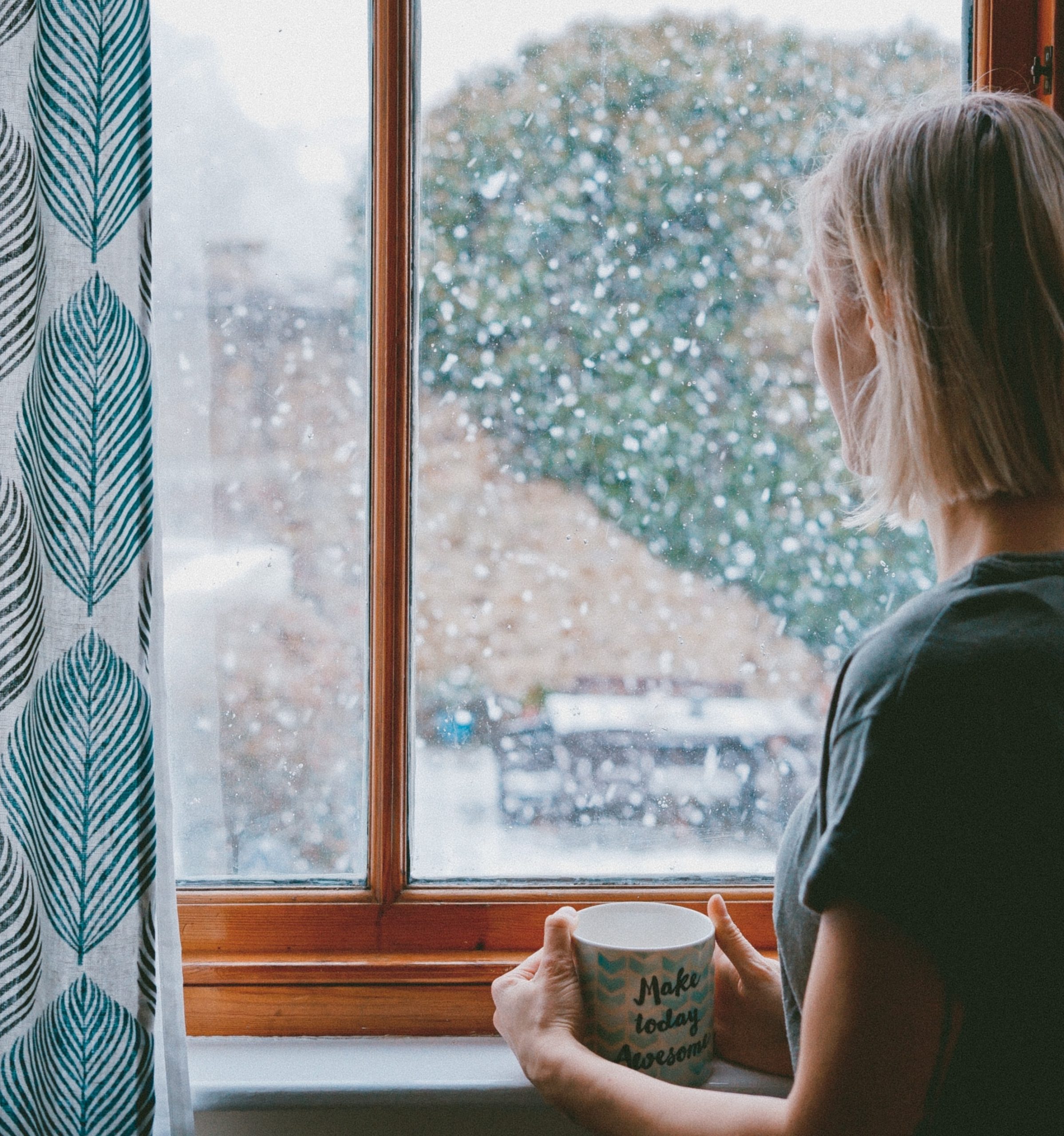 Woman holding a mug watches from a window as snow falls outside.