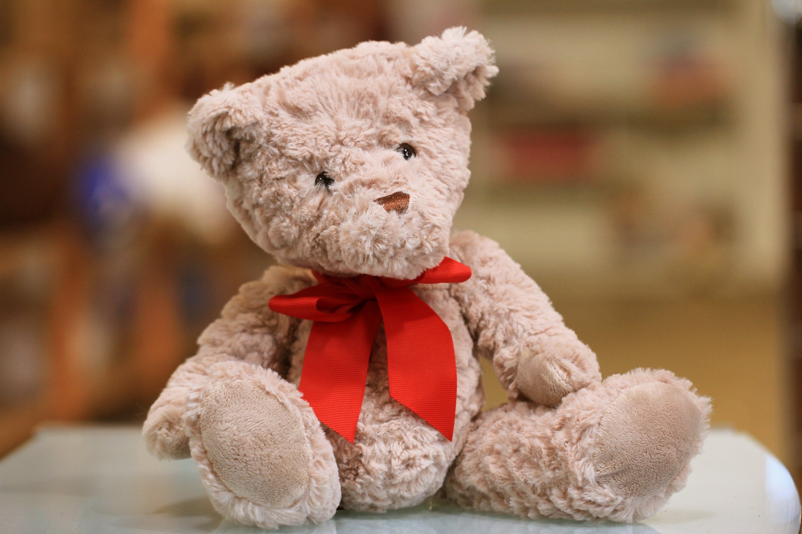 A fluffy tan teddy bear with a red bow around his neck sits on a table