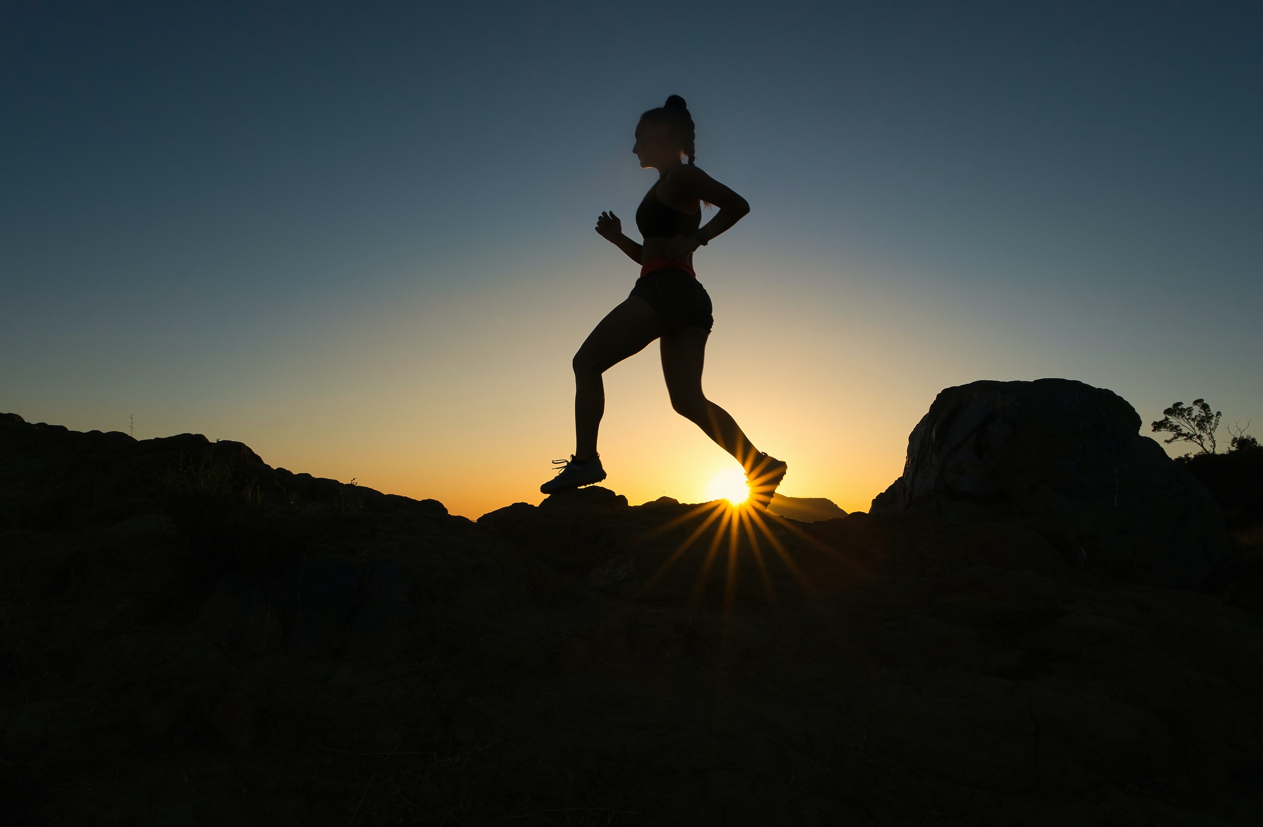 Silhouette of a woman runs across some rocks as the sun starts to rise behind her