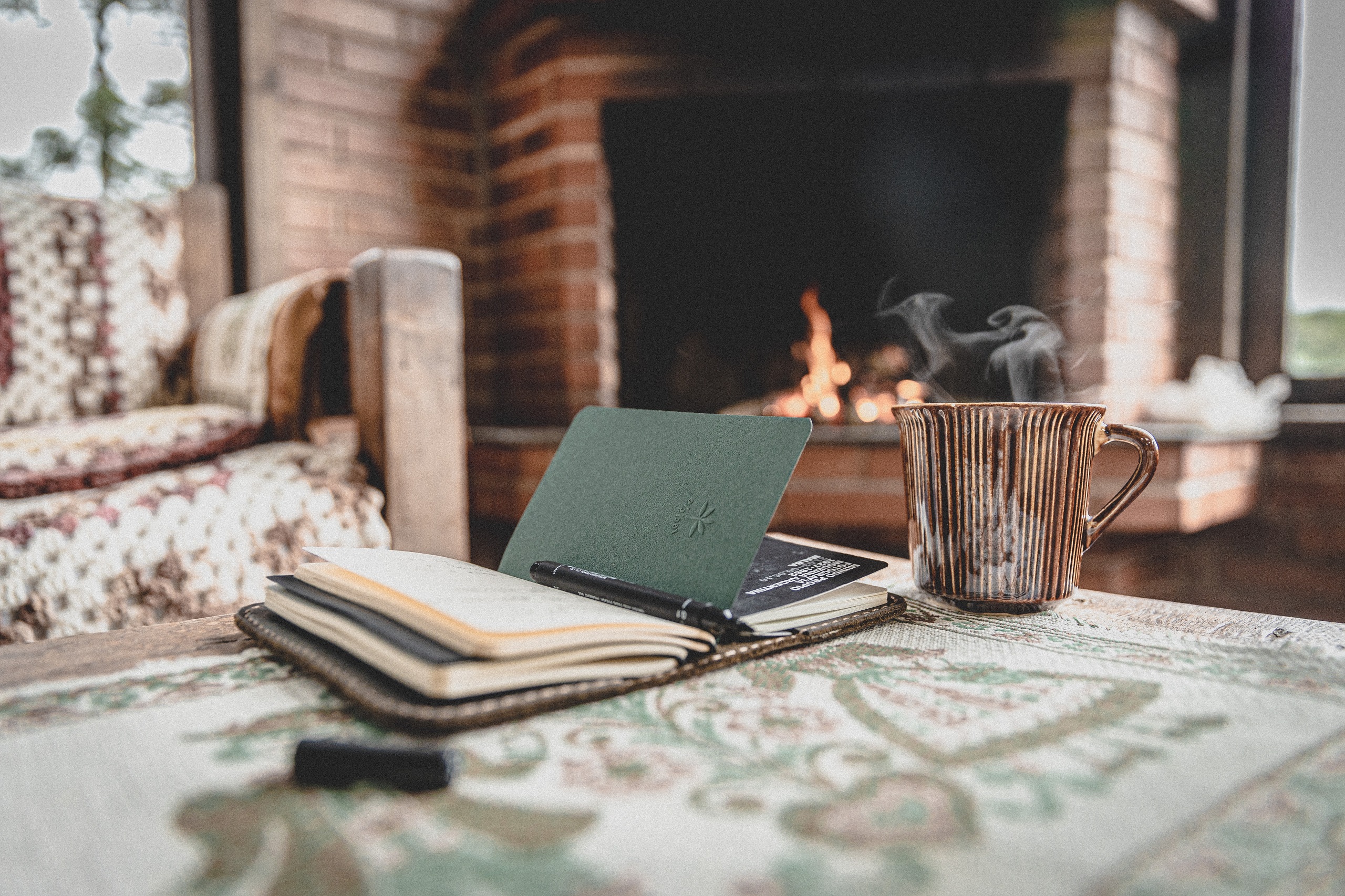 An open book sits on a table next to a steaming mug with a hot drink. In the background is an armchair and a lit fireplace.