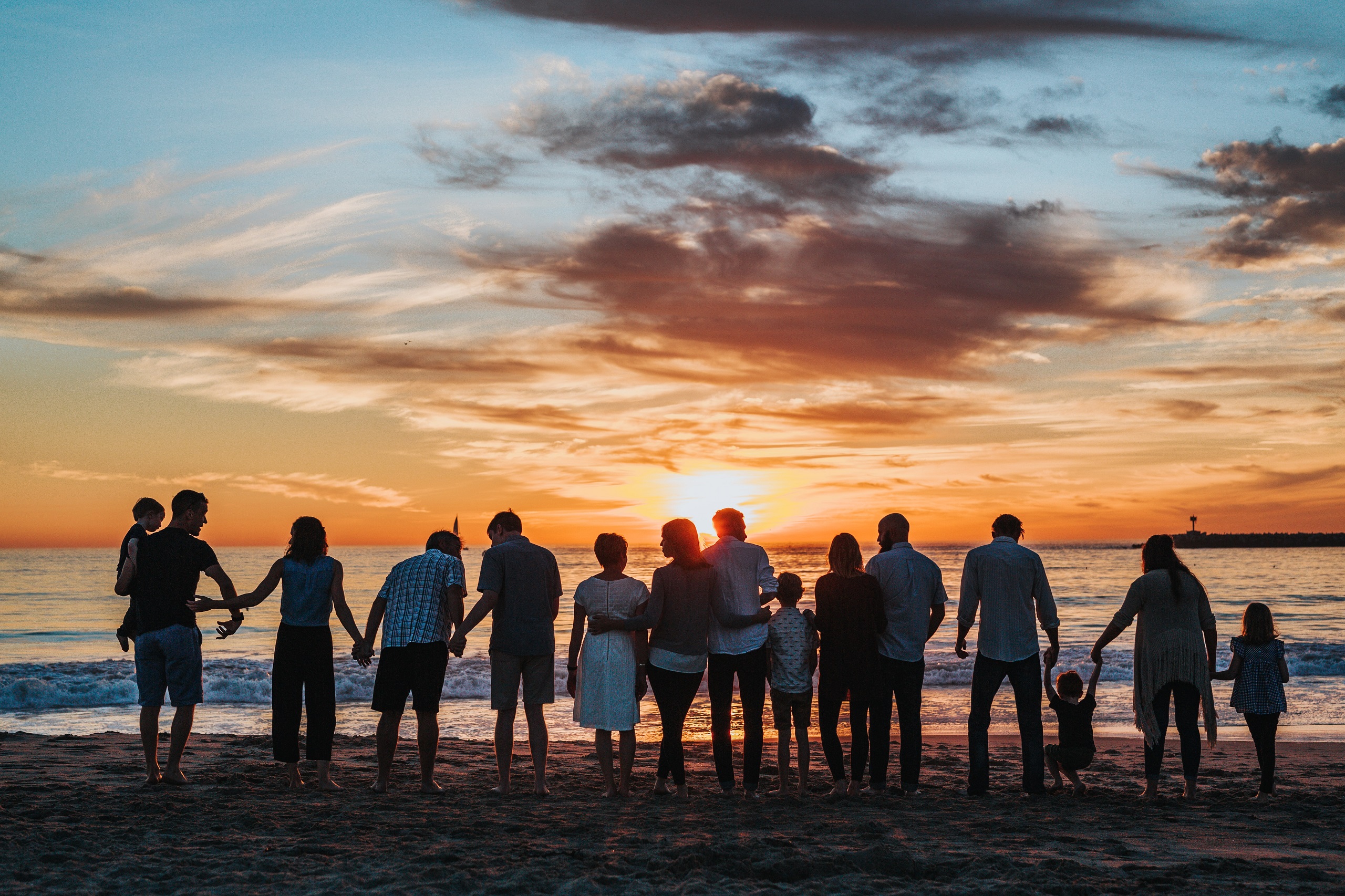 A large group of people of all ages stand on a beach facing the ocean at sunset