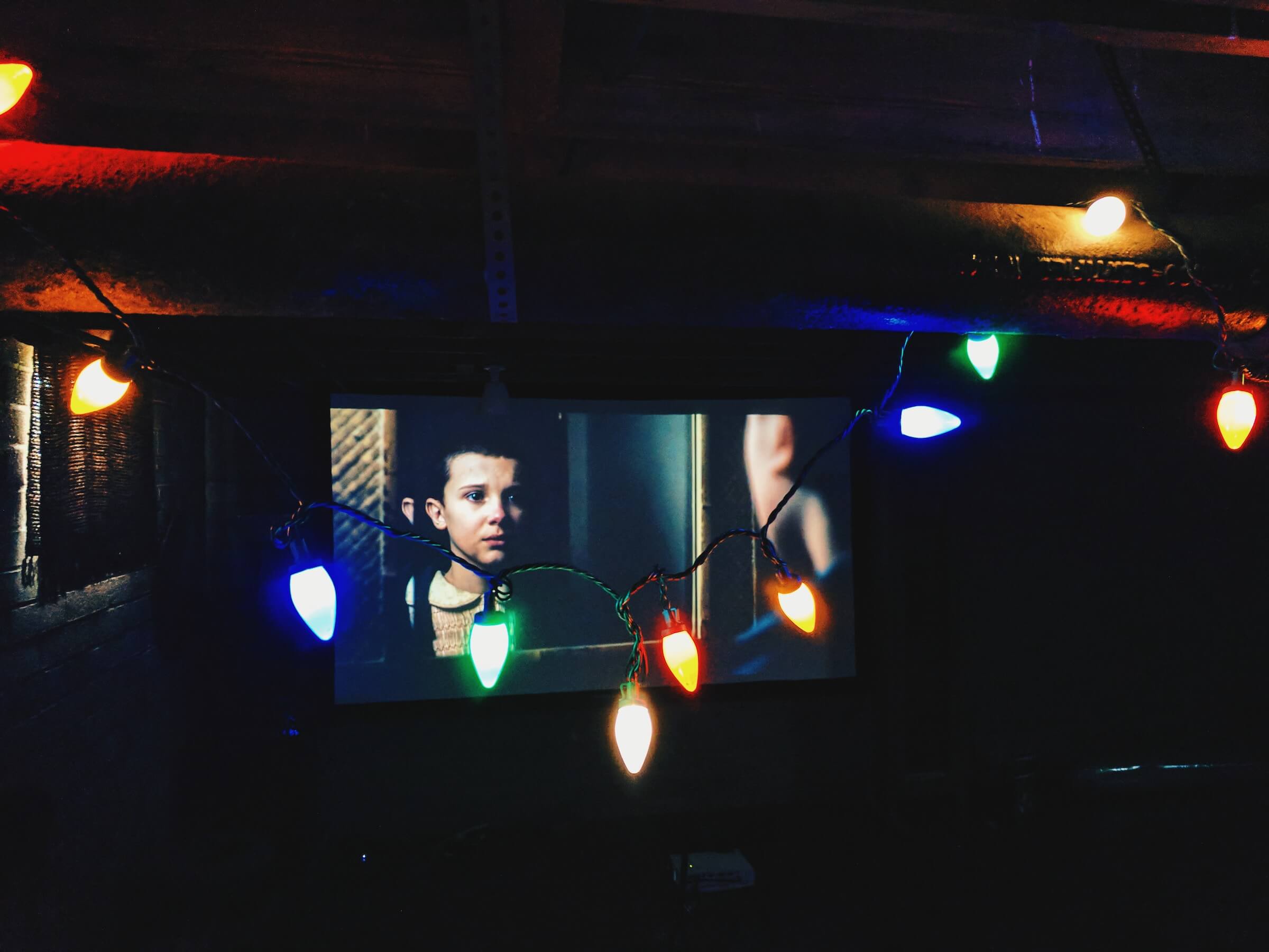 A scene from "Stranger Things" on a screen; a string of Christmas lights hang in front of the screen.