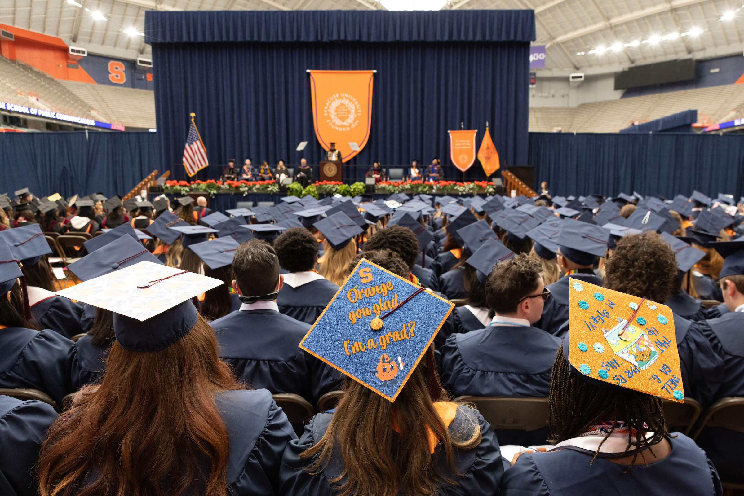 Students in graduation caps and gowns at Commencement. Writing on one of the caps is visible and says "Orange you glad I'm a grad?"