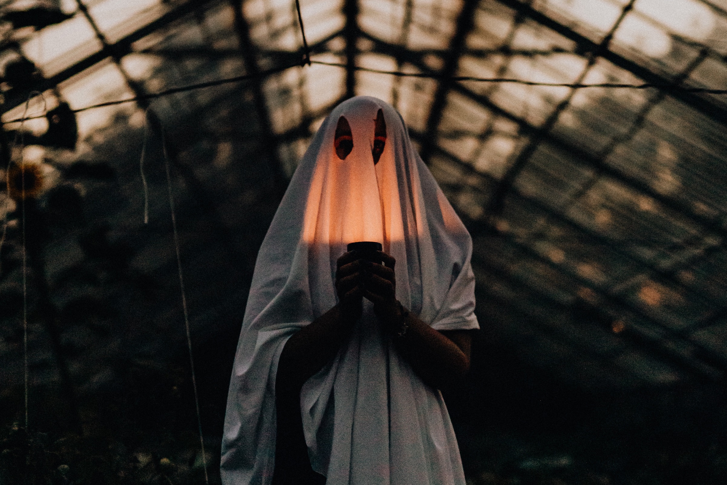 A person dressed as a ghost stands in a greenhouse with a candle illuminating their "face."