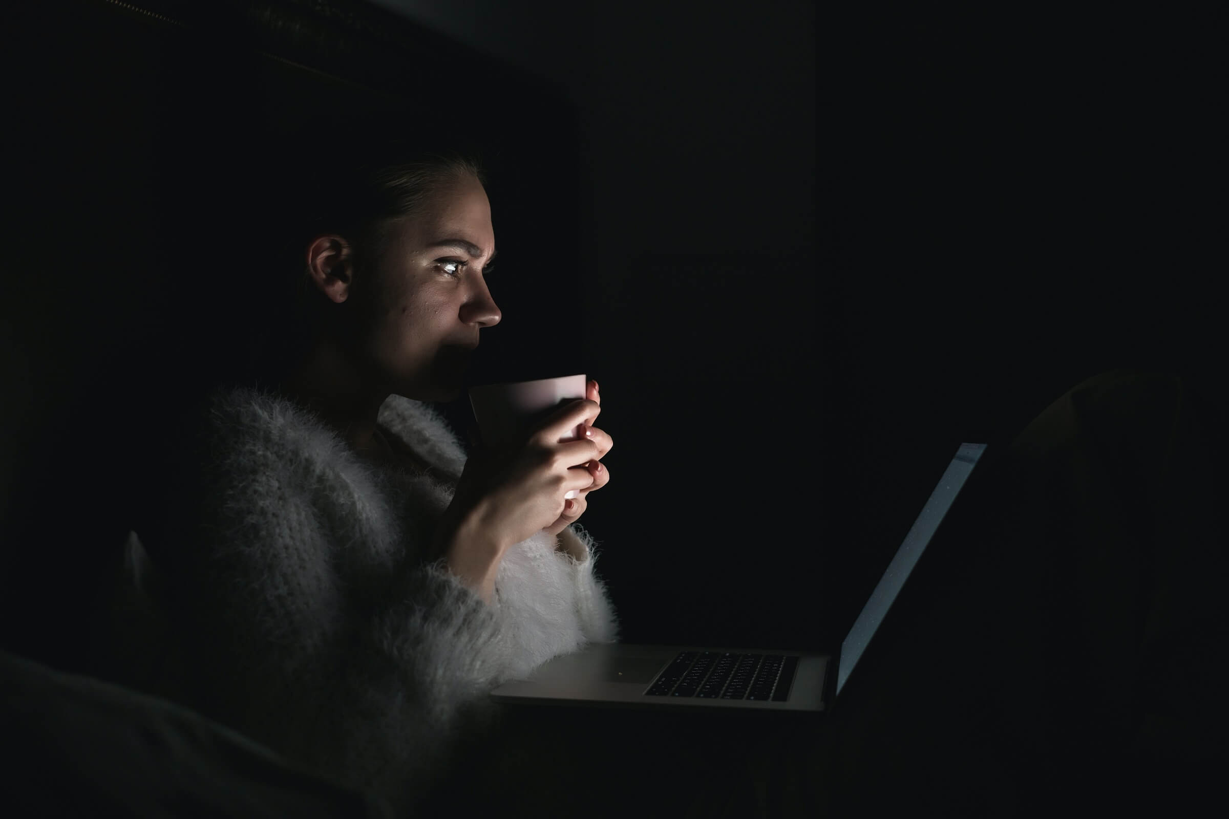 A woman holding a mug sits in the dark watching something on her laptop screen.