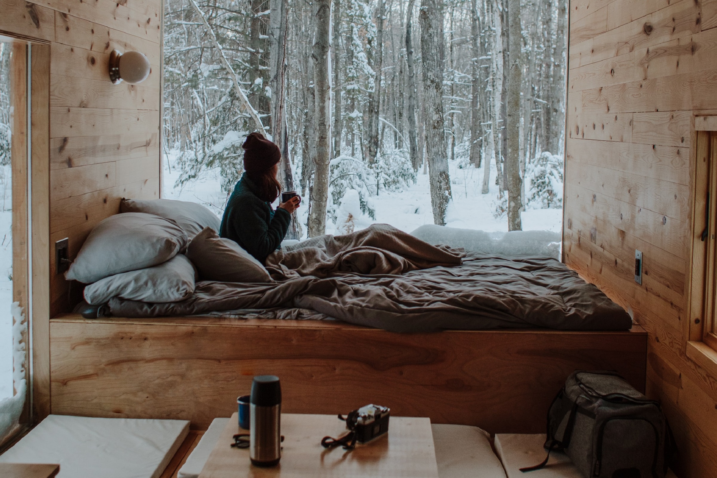 Person sits on a bed and looks out at snowy woods through a large window.