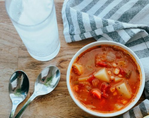 A bowl of minestrone soup on a wooden table. Beside it are two spoons, a glass of water, and a dishcloth