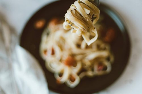 Camera focuses on a spoonful of noodles while a bowl of noodle soup is out of focus behind it.