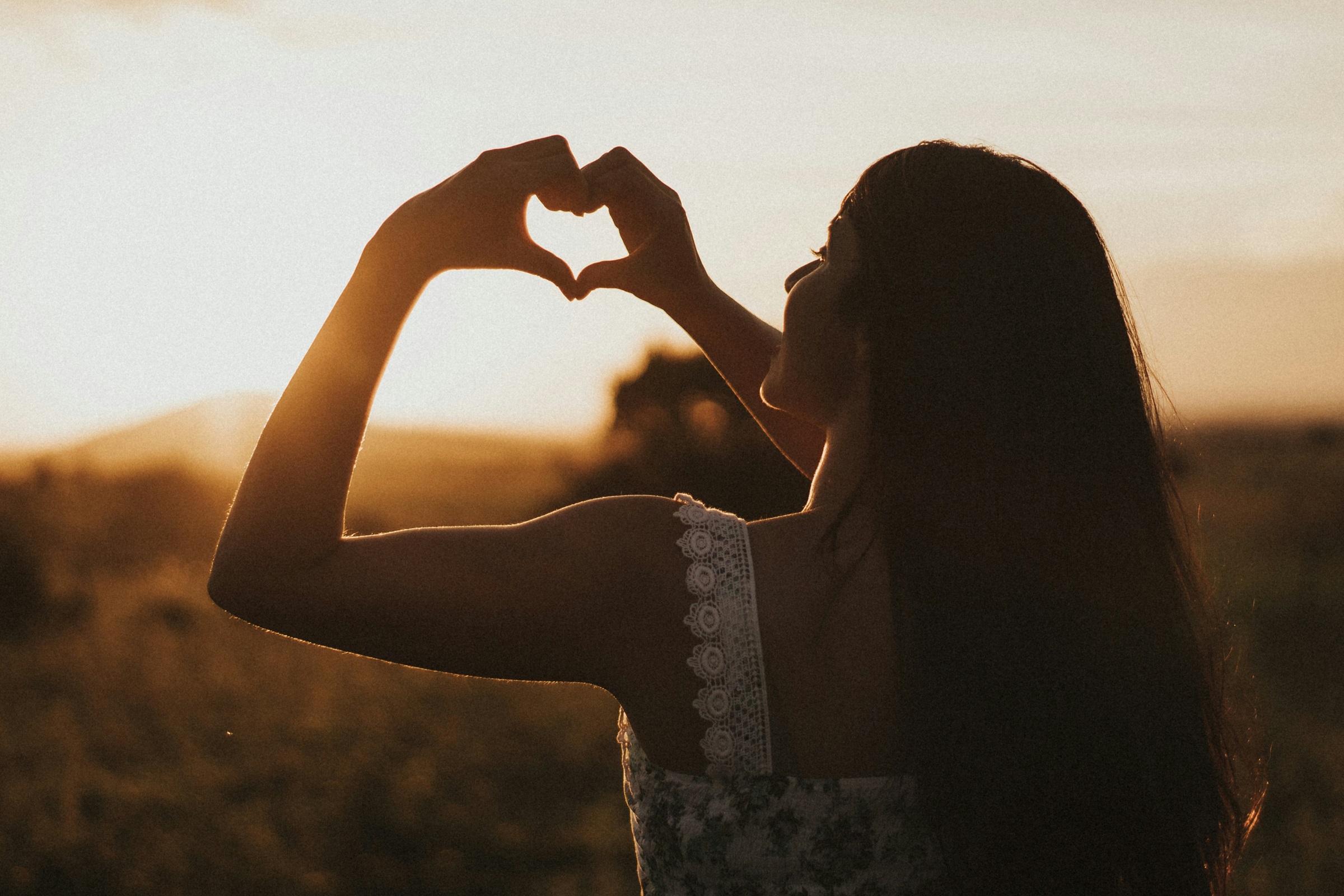 A woman makes a heart with her hands and holds it up to a sunset.