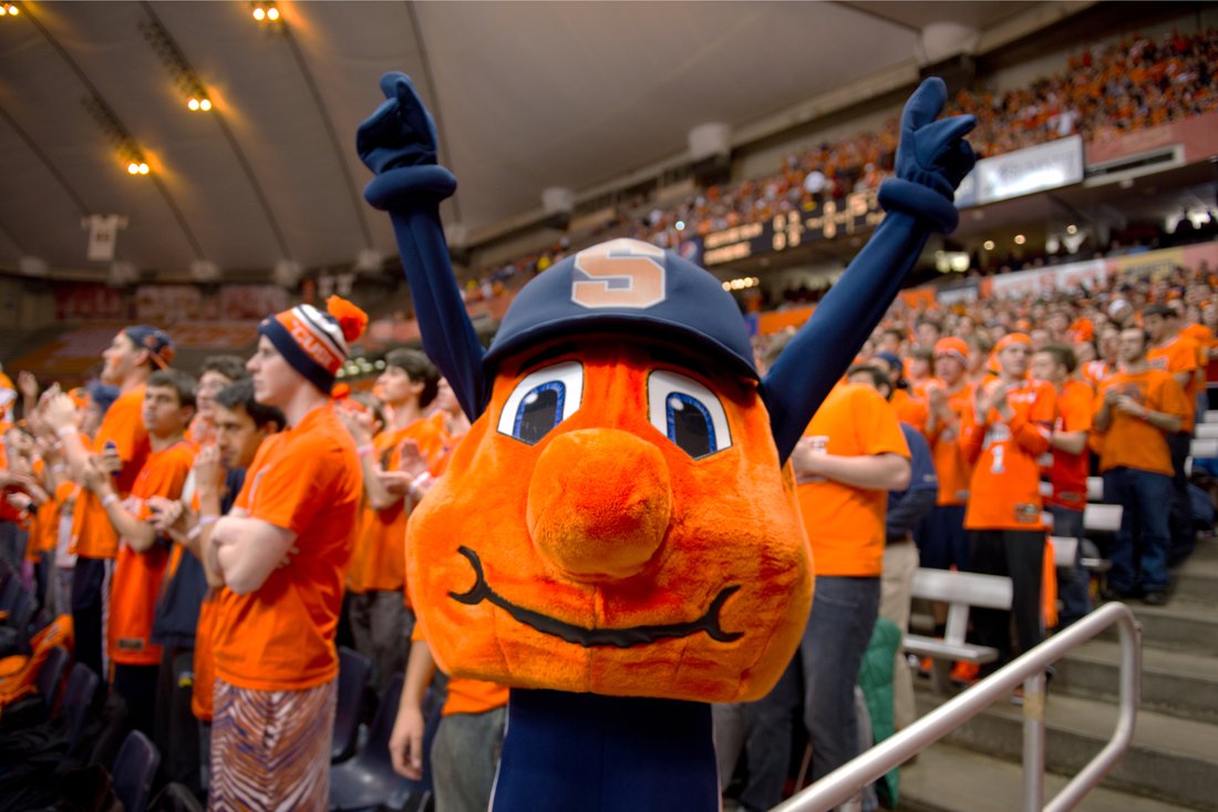 Otto the Orange stands with their hands raised in front of a crowd of students dressed in Syracuse colors at a game at The Dome.