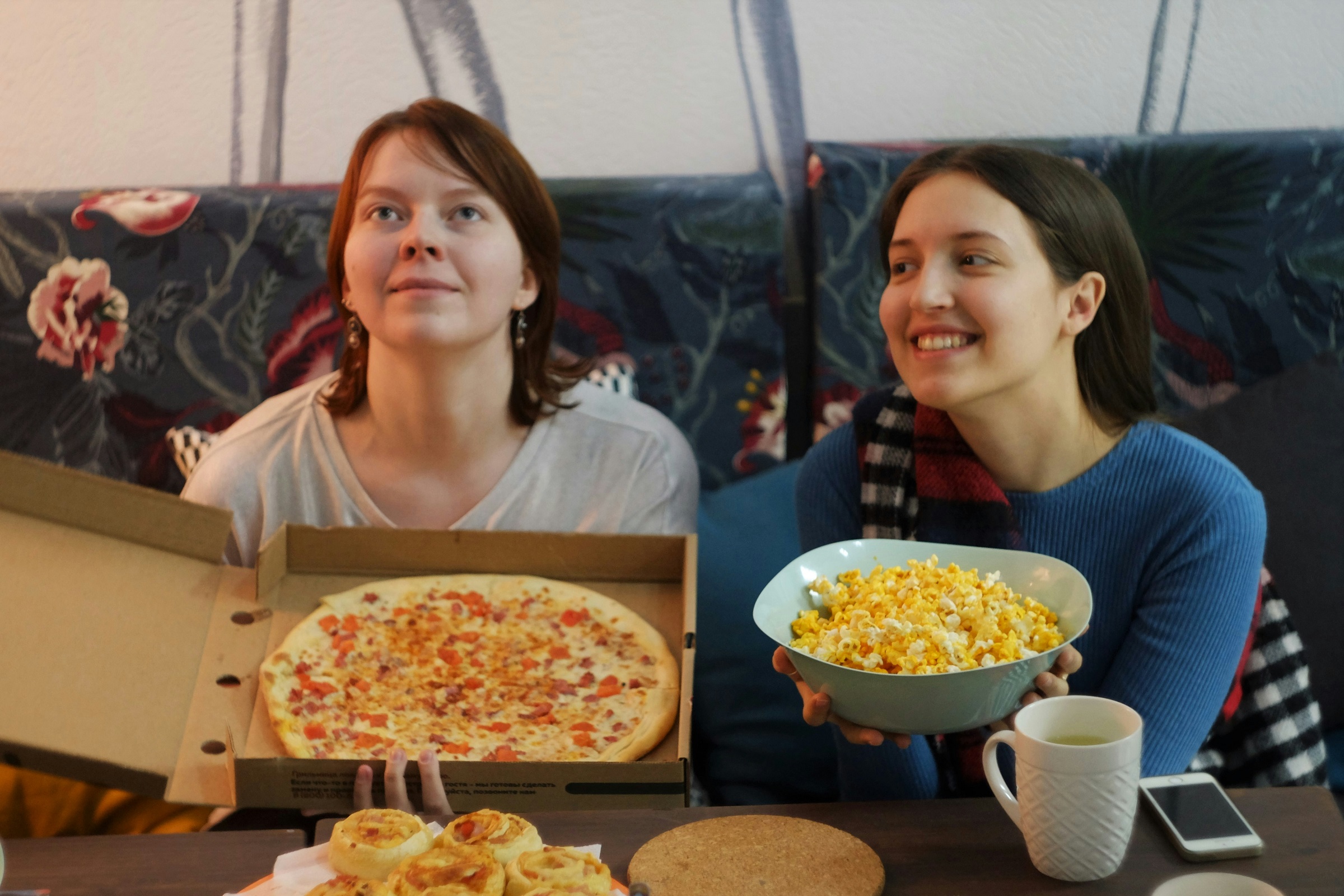 Two friends sit on a couch watching a movie. One holds a pizza box and the other holds a bowl of popcorn.
