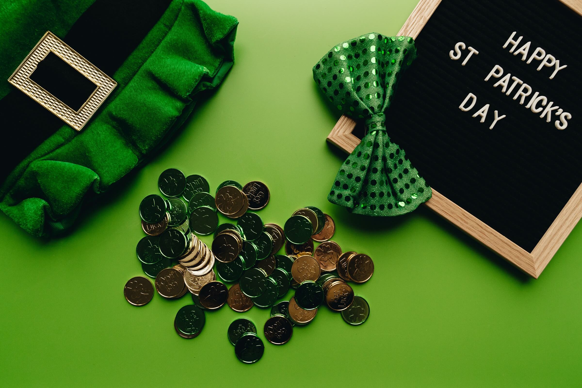 A pile of coins, a leprechaun hat, a green shiny bow and a memo board that says "Happy St. Patrick's Day" lay on a green backdrop.