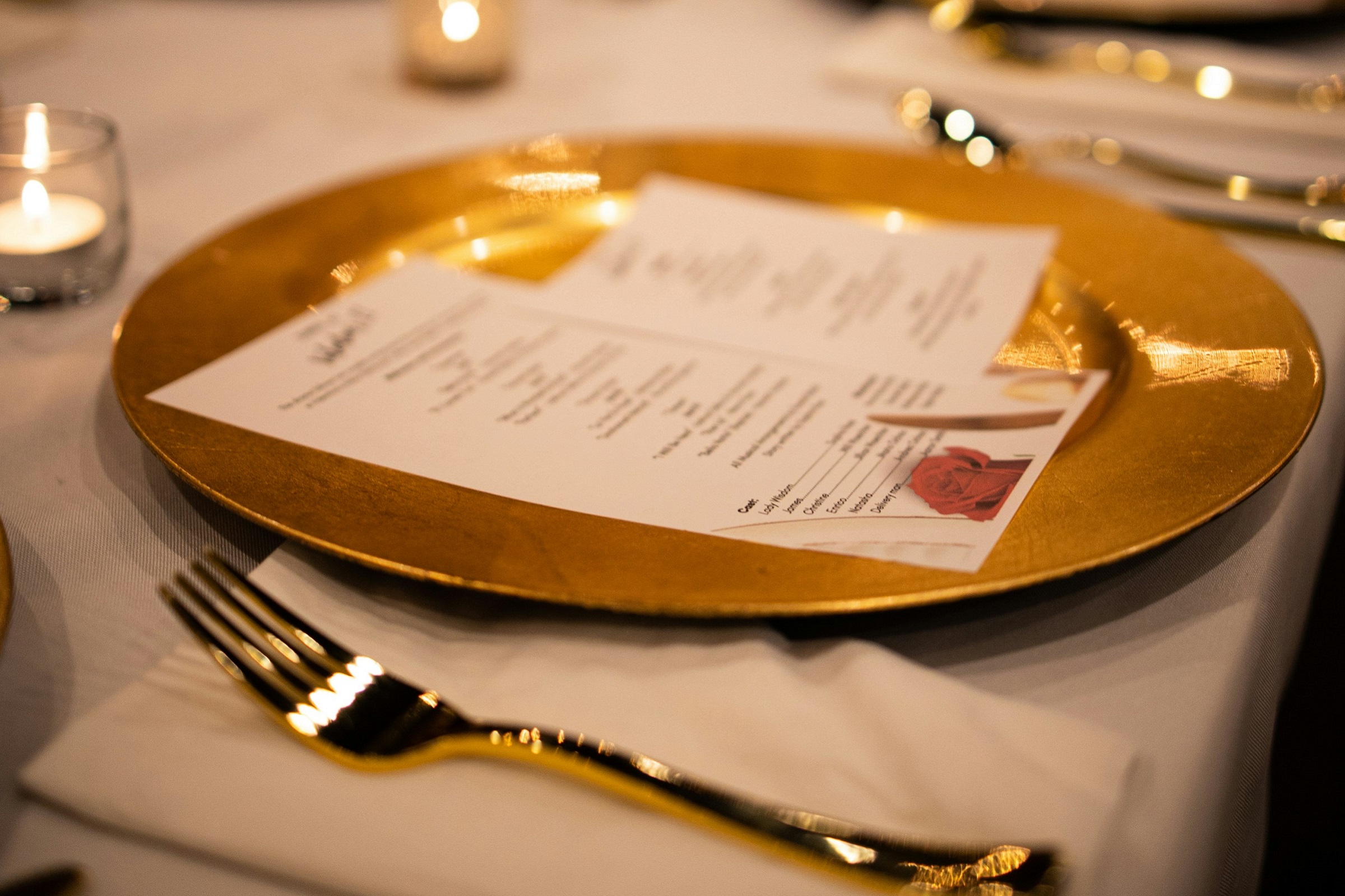 A paper menu rests atop a shiny gold dinner plate on a set table.