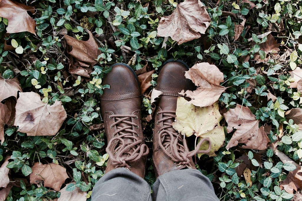 A person wearing boots takes a photo of their boots as they stand in grass scattered with fallen leaves.