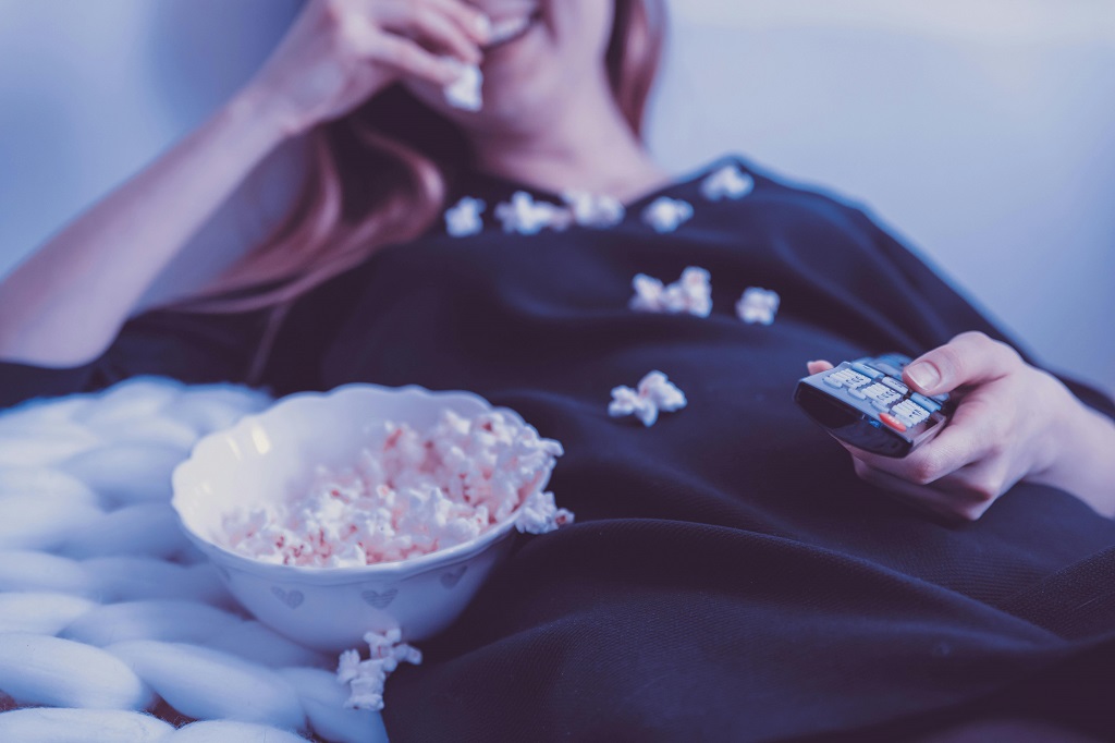 A woman lounges on a couch holding a TV remote and eating popcorn.