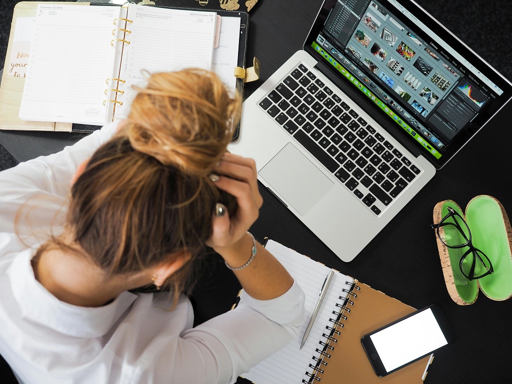 A woman surrounded by open notebooks and an open laptop holds her head in her hands.