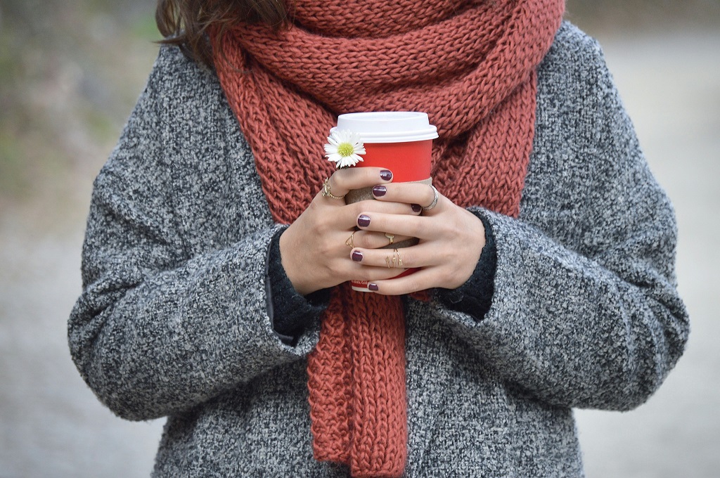 A woman in a sweater and a scarf holds a to-go coffee cup.