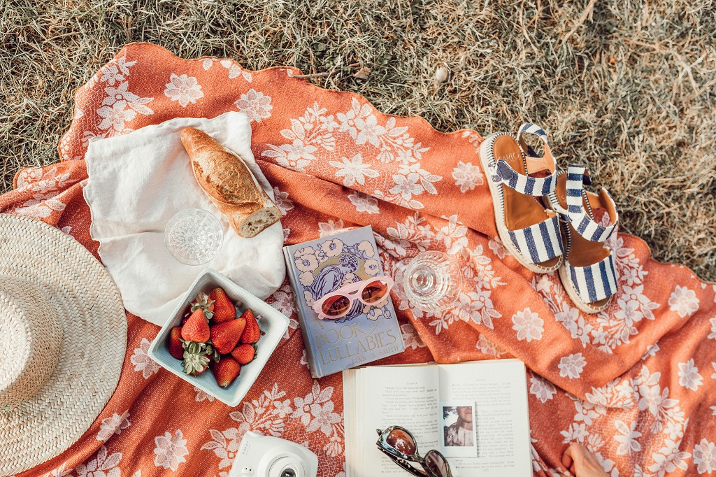 Bread, strawberries, books, a pair of sandals and a hat lay on top of a picnic blanket on the grass