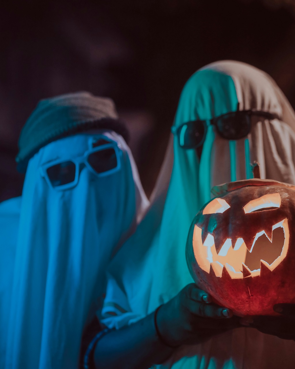 Two people in ghost costumes stand together for a photo with a jack-o-lantern