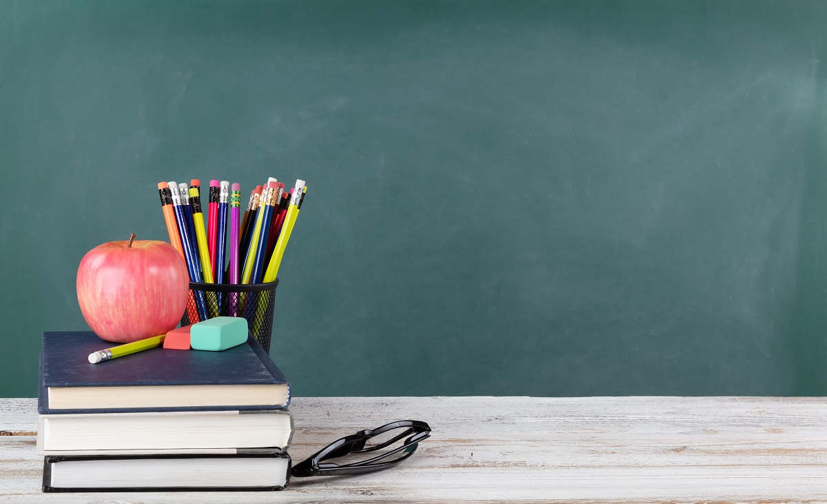 A pile of books and school supplies on a white desk in front of a chalkboard