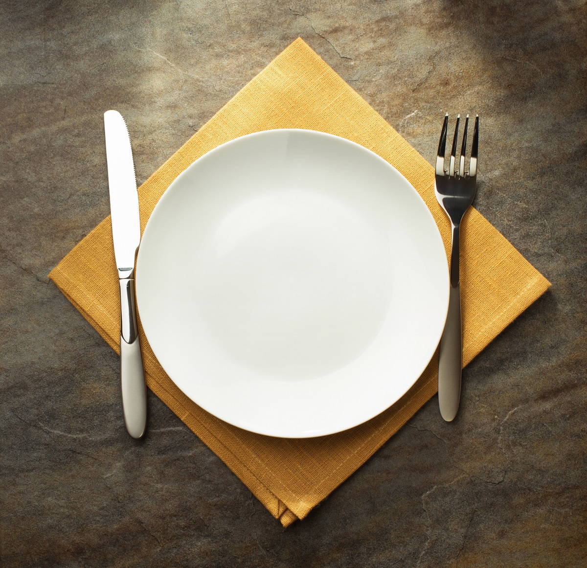 A dinner plate, knife and fork on top of a yellow cloth napkin on a wooden table.