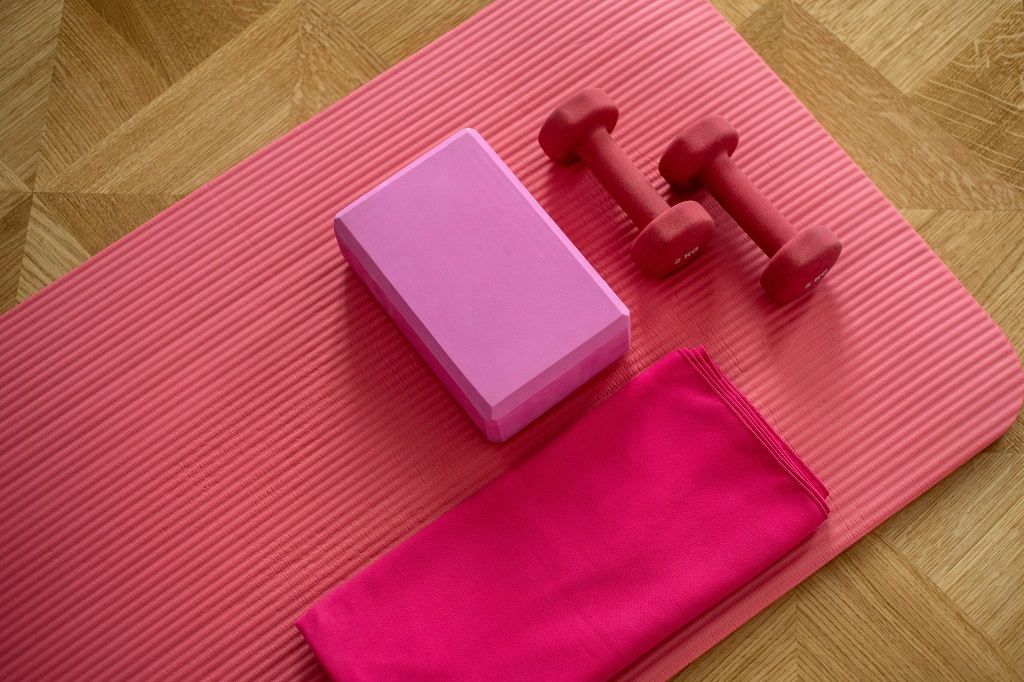 A pink yoga mat, pink block, pink weights, and a pink towel lay on a wooden floor.