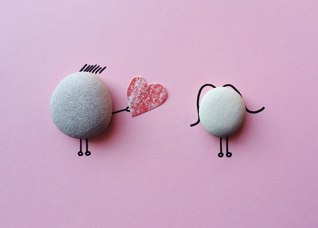 Two pebbles on a pink background. Hair, arms and legs were drawn on the pink background to make the pebbles look like people, and one of the pebbles is giving a construction-paper heart to the other.