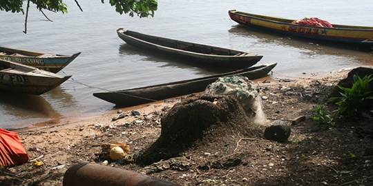 A canon relic rests on shore in Sierra Leone at the site of an 18th-century British slave trading fort, a research site for many years for Professor Christopher DeCorse. The boats are smaller versions of the one he fell from.