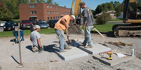 Workers prepare a bed of granite pavers to support the new memorial honoring those lost and injured in a barracks fire in Jan. 1959 on the Syracuse University campus. The seven tiles in the base will represent the seven airmen who died in the fire. The memorial is located on Lambreth Lane, South Campus.