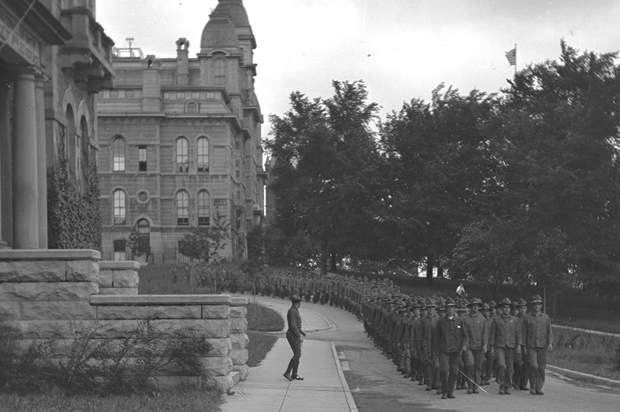 En esta foto, alrededor de 1918, el Cuerpo de Entrenamiento del Ejército de Estudiantes marcha en formación por el Salón de Idiomas. Esta es una de una colección de fotos de los Archivos de la Universidad de Syracuse que muestran la actividad de la Primera Guerra Mundial en el campus.