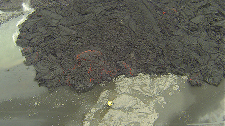 An overflight view of Jeffrey Karson, in yellow coat, shows him making measurements of the lava flow front. This scene of an active flow lobe on a riverbank is recorded by drone flight at about 100 meters altitude. Karson Earth is an Earth sciences professor in the College of Arts and Sciences regularly travels to Iceland to study volcanic structures.