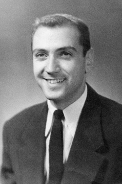 Black and white portrait of a smiling person in a dark suit and tie against a plain background
