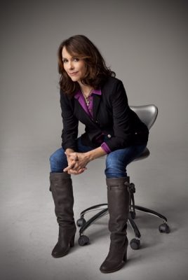 Person seated on a gray office chair wearing a black blazer with purple accents, purple shirt, jeans, and brown boots against a gray background.