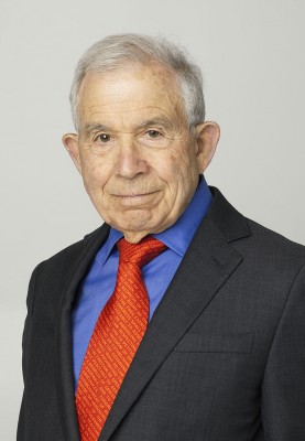 Person with light hair wearing a dark suit, blue shirt, and red tie against a plain light background.