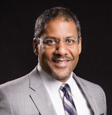 Smiling person in glasses wearing a light gray suit and striped tie against a dark background.
