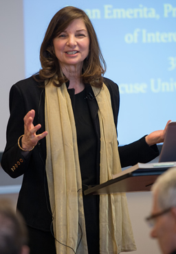 person delivering a presentation from a podium, gesturing with their hands, while a display screen is visible in the background