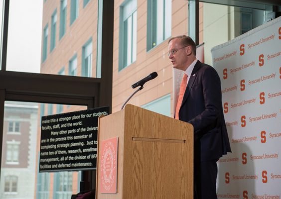 Chancellor Syverud speaking behind dais and against wall of windows